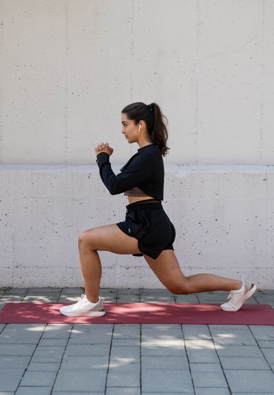 Conjunto atlético: camiseta negra de manga larga, sujetador deportivo gris, pantalones cortos negros. Zapatillas blancas. Realizando una zancada sobre una esterilla de yoga roja, fondo de hormigón gris.