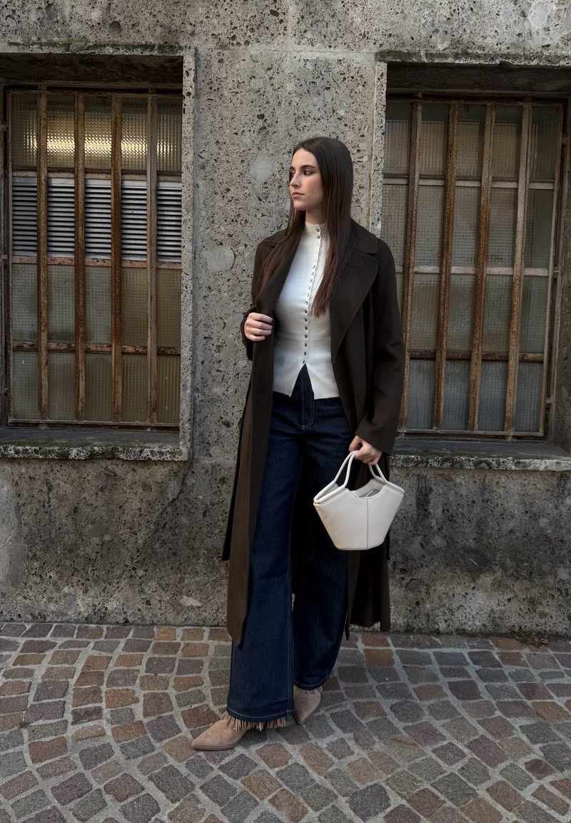 Femme en long manteau foncé, haut blanc boutonné, jeans à jambes larges, tenant un sac à main blanc, debout sur une rue pavée près d'un mur en pierre texturé.
