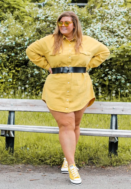 Jaune robe chemise boutonnée avec deux poches poitrine et une ceinture noire. Associée à des baskets Converse blanches avec des accents jaunes. Fond de feuillage vert.