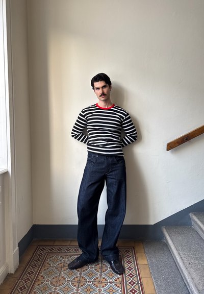 Hombre con bigote negro que lleva una camiseta a rayas blancas y negras con collar rojo, jeans anchos oscuros, de pie cerca de una escalera en un pasillo con azulejos.