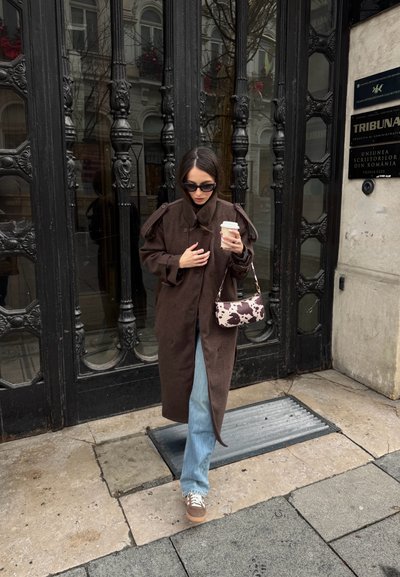 Mujer con abrigo marrón y vaqueros, sosteniendo una taza de café y una bolsa con estampado de vaca, caminando por la acera de la ciudad frente a una puerta negra ornamentada.