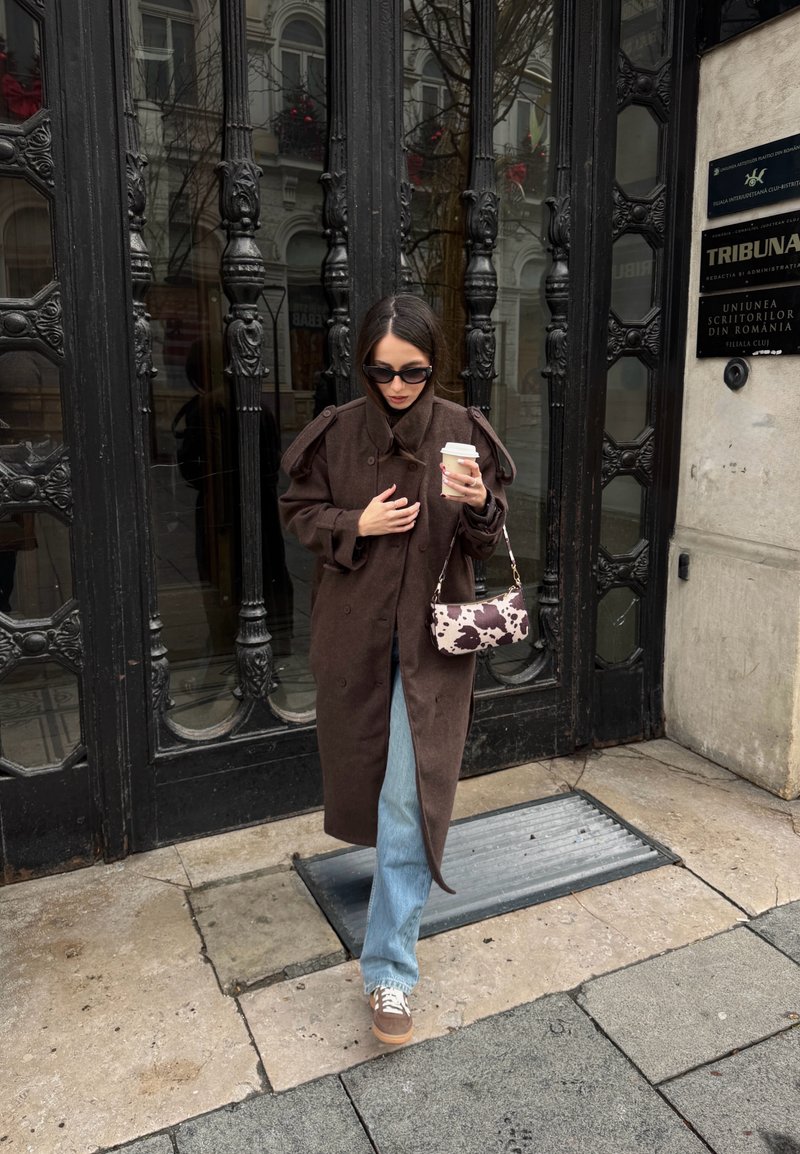 Femme en manteau marron et jean tenant une tasse de café et un sac imprimé vache, marchant sur le trottoir de la ville devant une porte noire ornée.