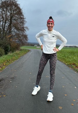 Chaqueta de forro polar blanca con cremallera y logo de North Face, leggings de estampado animal en rosa y negro, zapatos deportivos blancos, de pie en una carretera rural.