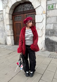 Veste rouge à fausse fourrure, haut rayé gris et noir, pantalon cargo noir, chaussures à plateforme noires, portant un sac à main noir avec des breloques.