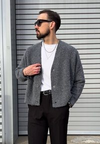 Siv, teksturiran kardigan z V-izrezom, zapenjanjem na gumbe in dolgimi rokavi, nošen prek bele majice in kombiniran s črnimi hlačami.