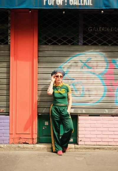 Chándal verde de Adidas con rayas amarillas, gafas de sol rojas y una banda para la cabeza estampada. El fondo presenta una tienda cerrada con graffiti colorido.