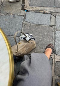 Bež usnjeni ročni torbici z belo in črno rutko, ob njej je rjava lakirana čevlja na tlaku iz kamenčkov. Ob njej leži siva tkanina.