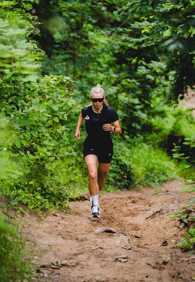 Zapatillas de correr en un sendero de arena; el sujeto lleva un atuendo negro con gafas de sol, rodeado de un exuberante follaje verde y un camino natural.