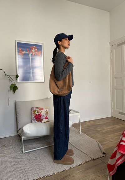 Bolso de mano de cuero marrón, suéter gris, pantalones de mezclilla oscuros, pantuflas marrones. El escenario presenta una pared clara, muebles blancos y una obra de arte floral.