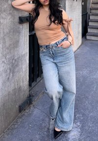 Beige fitted top dengan lengan ikat, dipadukan dengan celana jeans lebar berwarna biru muda yang dilengkapi dengan ikat pinggang rantai melingkar. Sepatu hak tinggi berujung runcing berwarna hitam.
