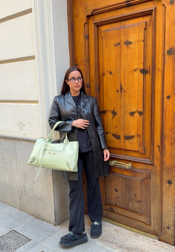 Kvinde med briller iført sort læderfrakke og bukser, holder en lysegrøn håndtaske og står ved en stor trædør.