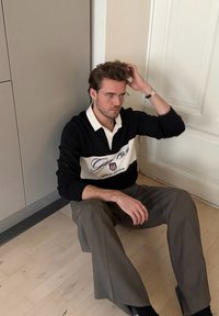 Pull-over rayé noir et blanc avec col, arborant un texte et un logo. Pantalon gris. Assis sur un sol en bois clair contre un mur.