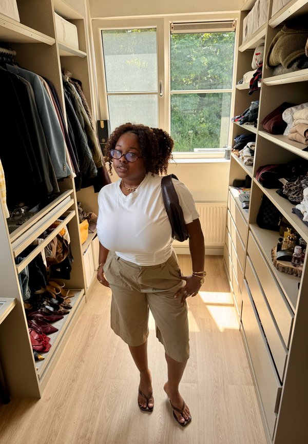 Mujer con pelo rizado, gafas, camiseta blanca, pantalones cortos beige y sandalias, de pie en un vestidor bien iluminado con estantes y ropa colgada.