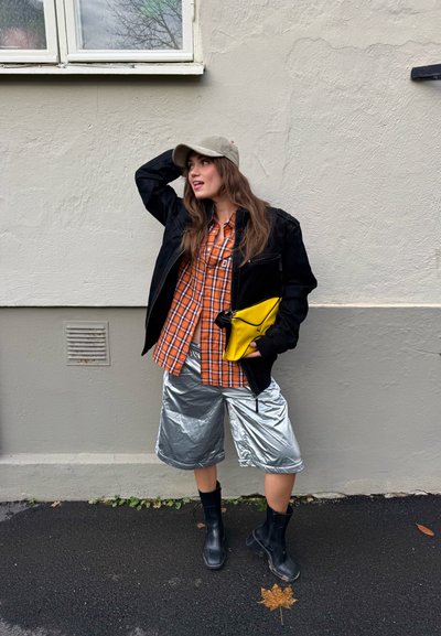 Chaqueta negra sobre una camisa de cuadros naranja, pantalones cortos plateados y botas negras. Sosteniendo una bolsa amarilla, de pie contra una pared lisa.