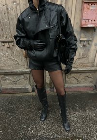 Personne portant une veste en cuir noire, un short à clous, des gants et des bottes hauteur genou, debout devant une vieille porte en bois usée.