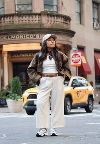 Veste en daim marron portée sur un haut blanc, pantalon beige à jambes larges, baskets noires et blanches, avec une casquette claire et une écharpe à motifs en accent.