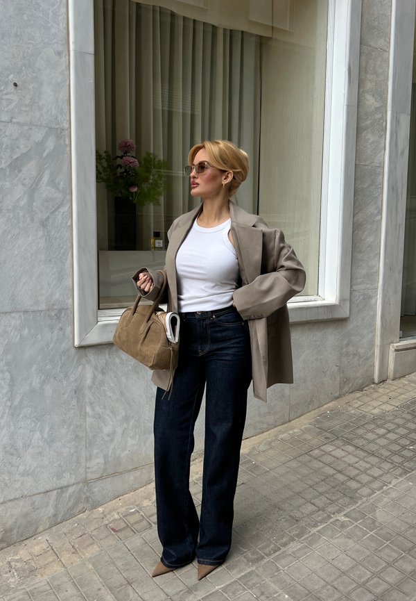 Femme portant des lunettes de soleil, un débardeur blanc, un blazer beige, un jean foncé et des chaussures pointues, tenant un sac à main beige et un journal près d'une fenêtre de bâtiment.