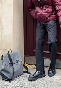 Mochila cinza com dois fechos frontais, alças pretas ajustáveis, superfície texturizada, colocada sobre paralelepípedos ao lado de uma pessoa com calças e botas pretas.