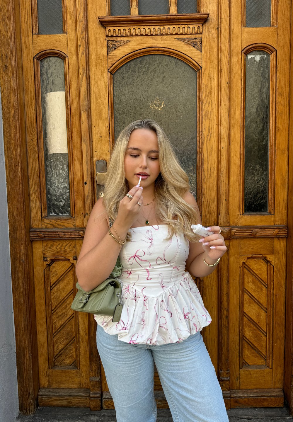 Weißes Oberteil mit rosa Schleifenmuster, Peplum-Saum; hellblaue weite Jeans; grüne Handtasche; hält Lipgloss; hölzerner Tür-Hintergrund.