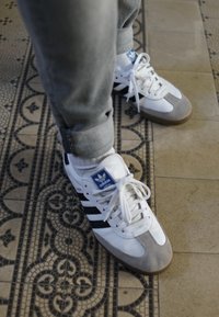 Sneakers brancos com acabamentos em camurça cinza, três riscas pretas e um patch de logótipo azul. Usados com jeans cinzas claros e meias brancas.