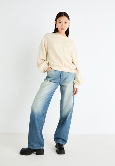 Sudadera beige con mangas abullonadas, combinada con jeans de pierna ancha de color azul claro con un lavado descolorido. Botas negras de tacón grueso completan el look.