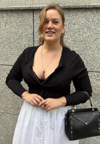 Mujer sonriendo frente a una pared de granito, vestida con un cárdigan negro sobre una blusa burdeos, falda blanca, anillo rojo, collar de oro y sosteniendo un bolso negro.