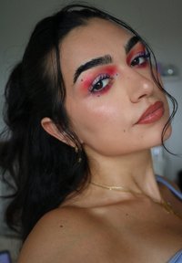Porträt einer Person mit dunklen Haaren, kräftigem roten und blauen Augen-Make-up, nude Lippenstift, goldener Schmuck und weicher Hauttextur vor einem neutralen Hintergrund.