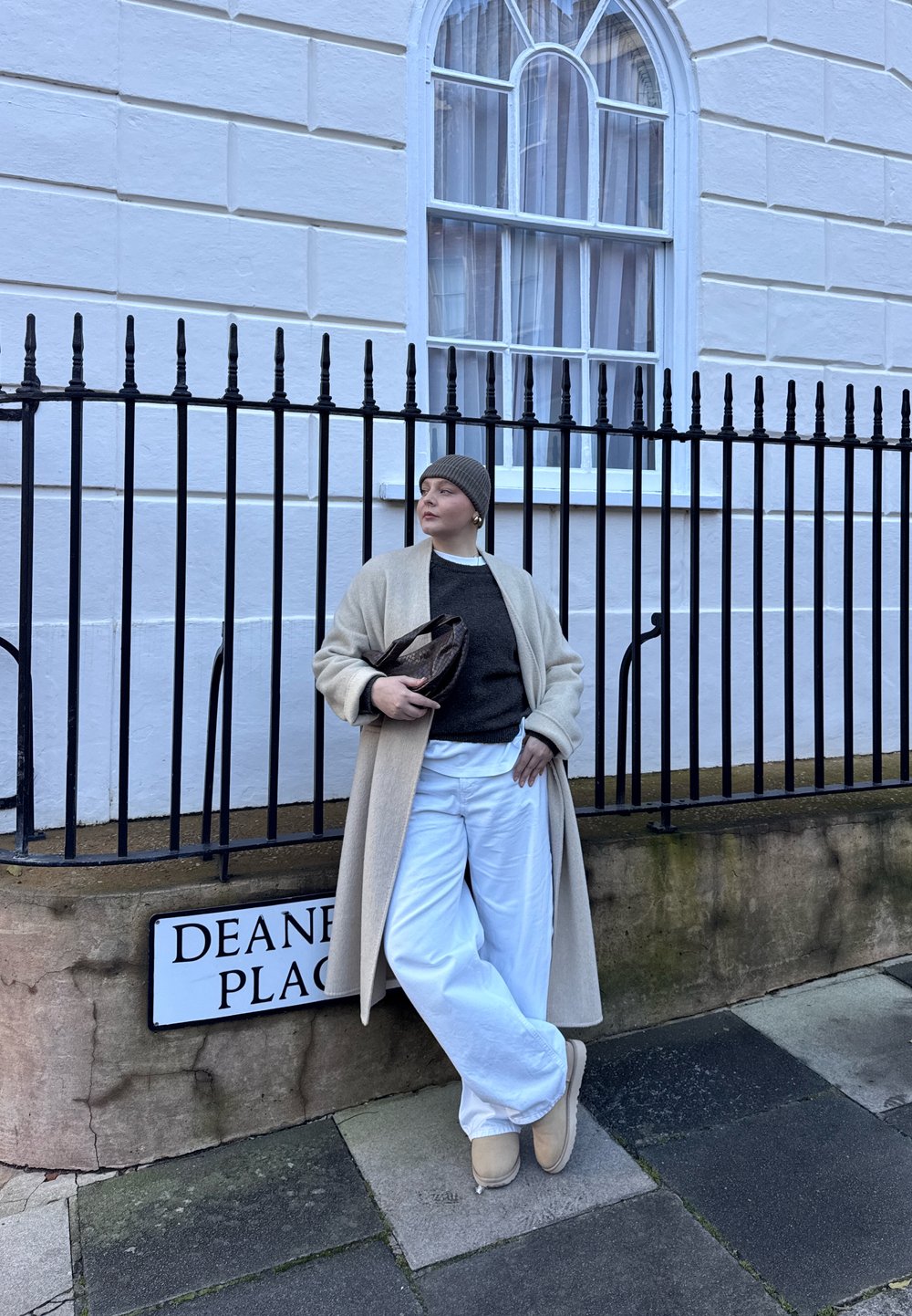 Personne en manteau beige, pull gris, pantalon blanc et bonnet appuyée contre une rambarde près d'un panneau de rue Deane Place.