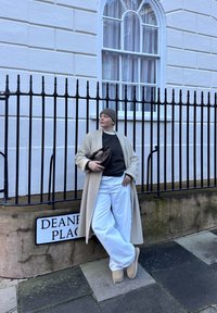 Personne en manteau beige, pull gris, pantalon blanc et bonnet appuyée contre une rambarde près d'un panneau de rue Deane Place.