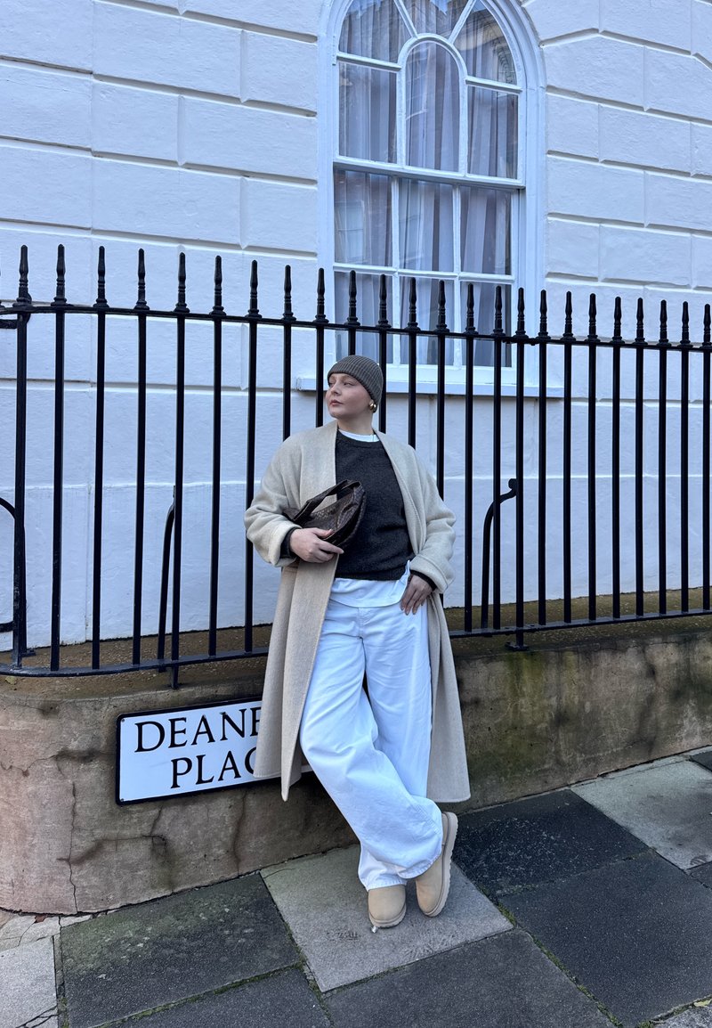 Personne en manteau beige, pull gris, pantalon blanc et bonnet appuyée contre une rambarde près d'un panneau de rue Deane Place.