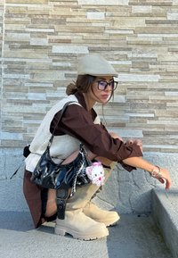 Gorra beis ajustada, camisa marrón de botones oversize, chaleco de punto crema, bolso negro brillante con llavero, botas beige hasta el tobillo con suelas texturizadas.