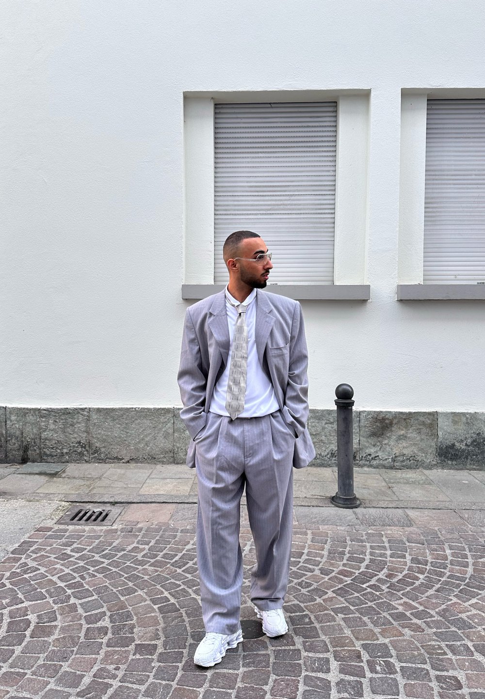 Ανοιχτό γκρι κοστούμι με ριγέ σχέδιο και φαρδιές πέτες, λευκό πουκάμισο και πουκάμισο με σχέδιο. Λευκά αθλητικά παπούτσια. Αστική περιοχή με πέτρινη επιφάνεια.