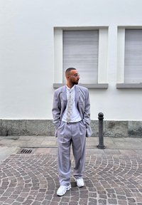 Ανοιχτό γκρι κοστούμι με ριγέ σχέδιο και φαρδιές πέτες, λευκό πουκάμισο και πουκάμισο με σχέδιο. Λευκά αθλητικά παπούτσια. Αστική περιοχή με πέτρινη επιφάνεια.