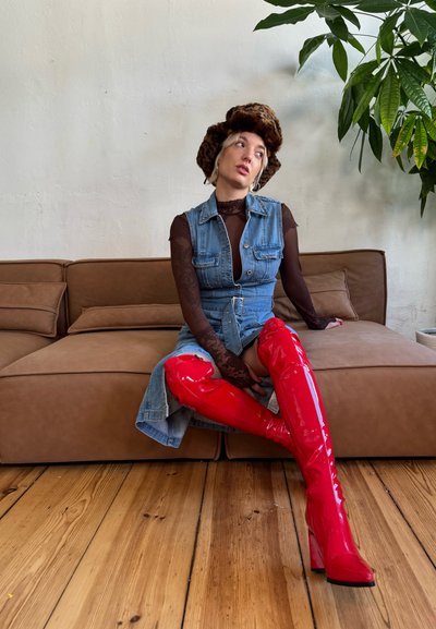 Modelo con un vestido de mezclilla, una blusa negra de manga larga transparente y botas de charol rojas hasta el muslo, sentada en un sofá marrón. Planta verde al fondo.