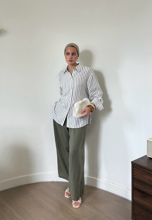 Femme portant un foulard beige, une chemise rayée blanche et noire, un pantalon large vert olive, tenant une pochette blanche, debout près d'une armoire en bois.