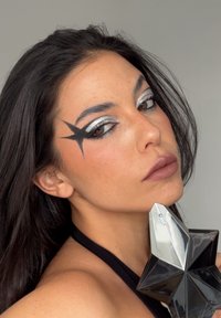 Svart parfymflaska i stjärnform med silverkork; djärv grafisk ögonmakeup i silver och svart; slät, strålande hudstruktur.