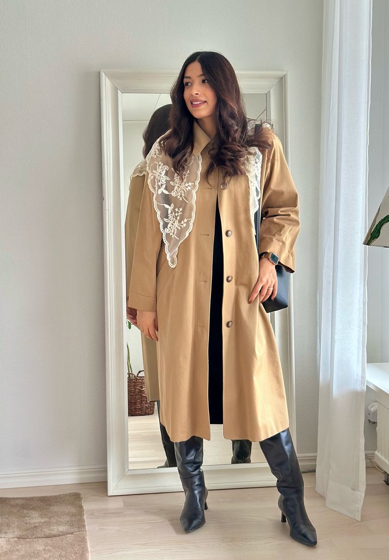 Mujer de cabello largo y oscuro, con abrigo beige, bufanda de encaje, vestido negro y botas negras hasta la rodilla, de pie frente a un espejo de cuerpo entero.