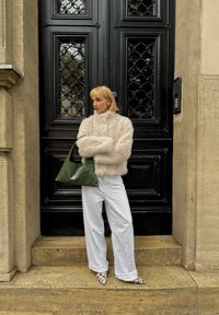 Femme blonde en veste blanche en fourrure et large pantalon blanc se tient sur des marches en pierre, tenant un sac à main vert devant une porte noire ornée.