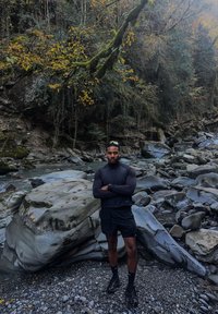 Atleet in donkere, nauwsluitende buitenkleding staat bij een rotsachtige rivieroevers. Natuurlijke elementen omvatten gladde, grijze stenen en herfstbladeren op de achtergrond.