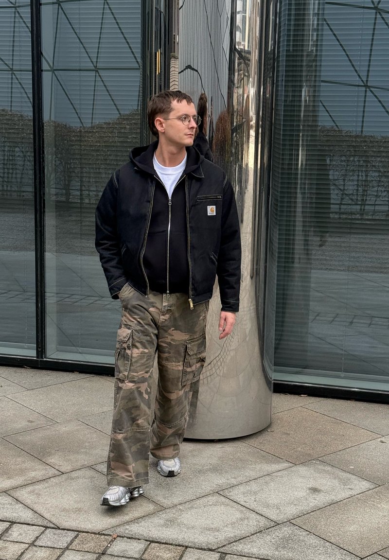 Homme portant des lunettes, une veste noire, des baskets blanches et un pantalon cargo camouflage marchant à côté d'une colonne métallique réfléchissante près d'un bâtiment en verre.