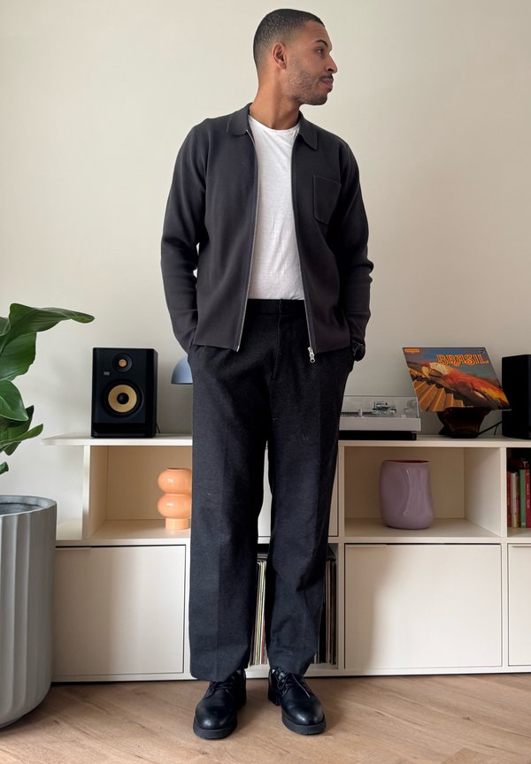 Mann in schwarzer Jacke, weißem T-Shirt und dunkler Hose steht mit den Händen in den Taschen in einem modernen Raum mit Lautsprechern und Vinyl-Schallplatten.