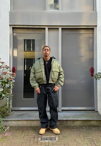 Giacca bomber verde con due tasche frontali, felpa nera, pantaloni in denim scuro e zoccoli beige, in piedi di fronte a una porta grigia.