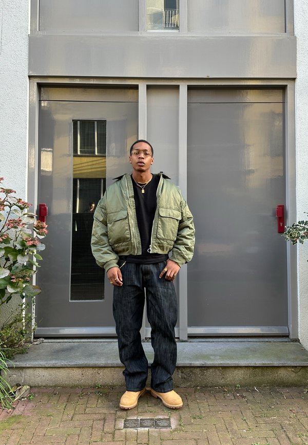 Grön bomberjacka med två framfickor, svart sweatshirts, mörka jeans och beige träskor, står framför en grå dörr.
