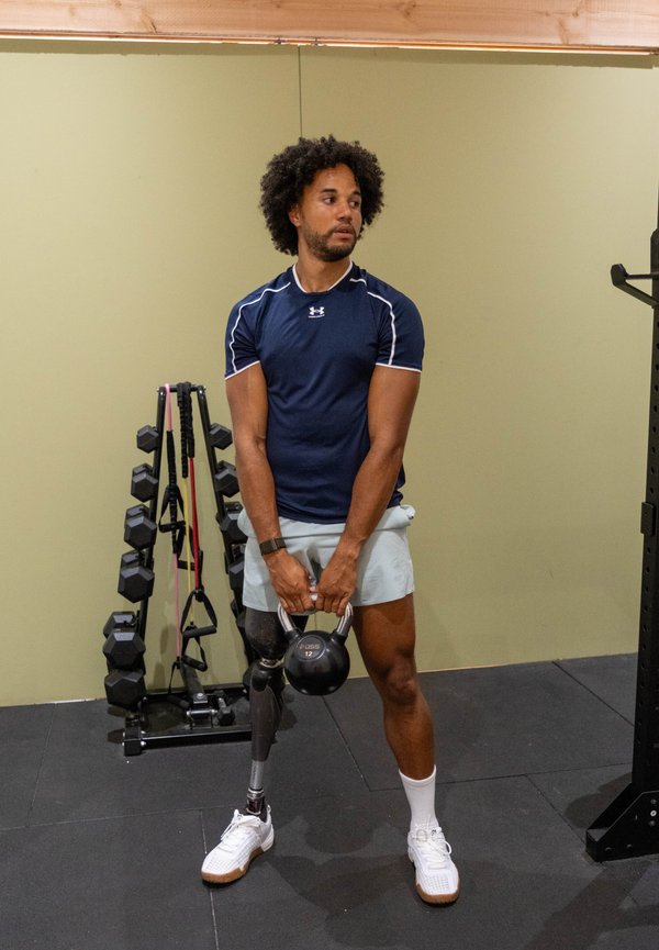 Uomo con gamba protesica che tiene un kettlebell in palestra, in piedi vicino a pesi e bande elastiche contro una parete beige.