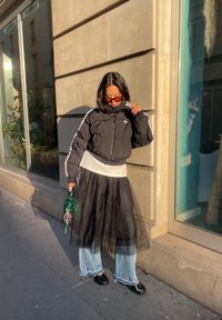Personne portant une doudoune noire, jupe en tulle noir superposée sur un jean bleu, chaussures noires, lunettes de soleil orange, tenant un sac à main vert sur un trottoir ensoleillé.