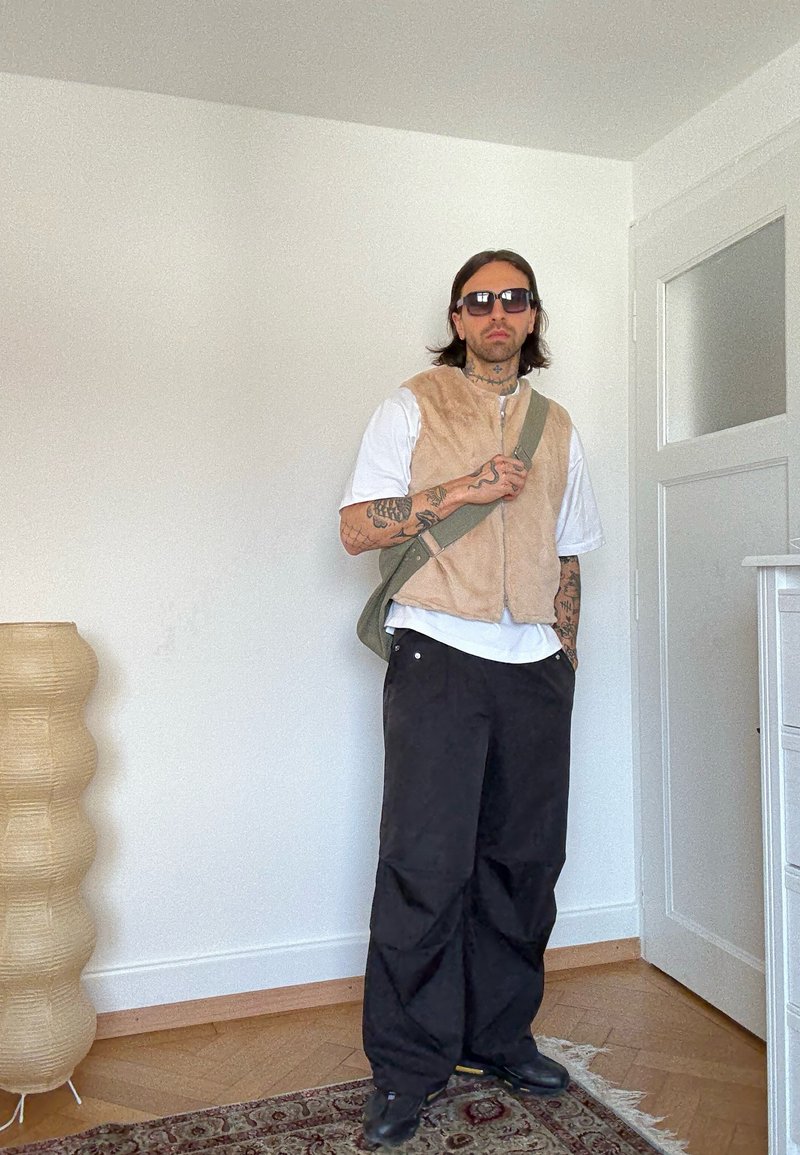 Man met tatoeages, zonnebril, beige vest over een wit T-shirt, zwarte wijde broek, staand in een kamer met houten vloer en een lamp.