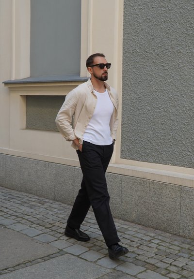 Camisa de botón de color beige claro sobre una camiseta blanca, combinada con pantalones negros y zapatos negros. Caminando por un camino de adoquines cerca de una pared texturizada.