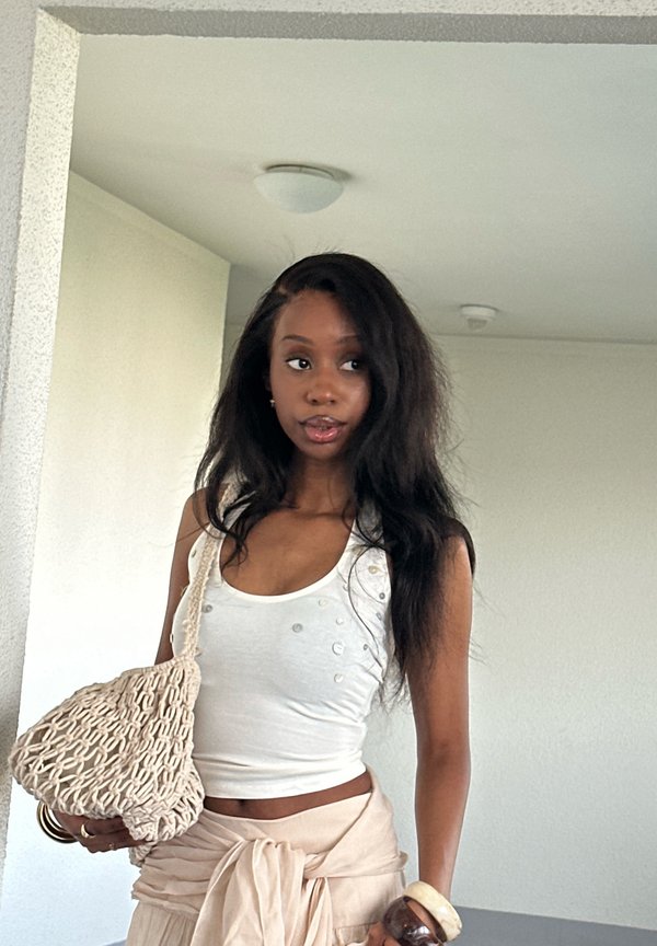Joven mujer en interiores con una camiseta sin mangas blanca y una falda beige cruzada, sosteniendo un bolso beige de punto sobre su hombro.