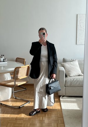 Blazer negro ajustado, camiseta blanca, falda larga crema, sandalias negras; sosteniendo un bolso estructurado negro. Suelo de madera, interior moderno.