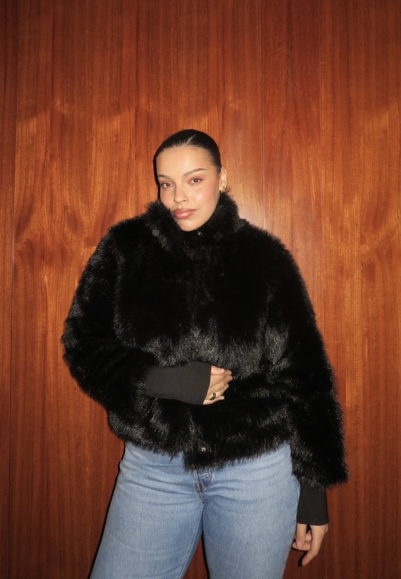 Ung kvinne med bakoverstrøket hår iført svart fuskepelsjakke og lyseblå jeans, står foran en bakgrunn av trepanel.